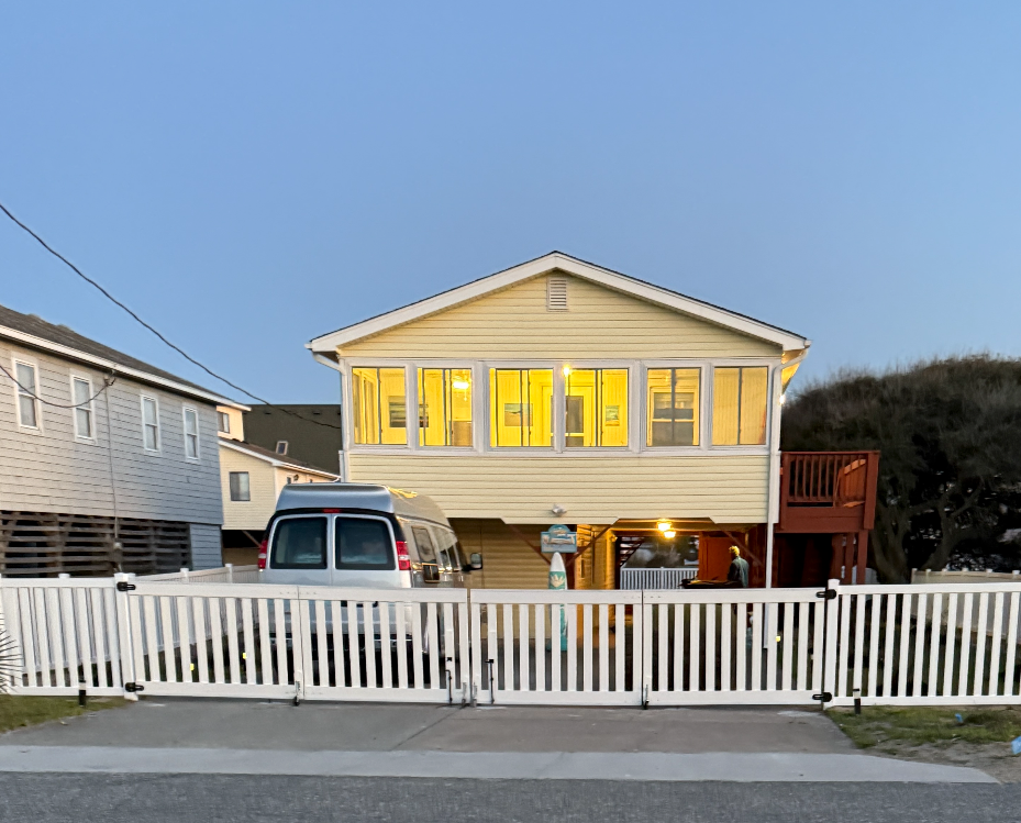 Yellow beach house with a white fence, a van parked underneath, and lit windows.