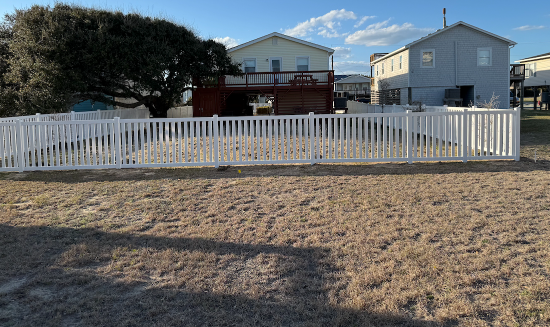 White picket fence encloses yard, houses in background, sunny day.