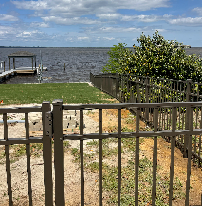 Brown fence on a grassy waterfront, with a dock in the distance. Overcast sky.