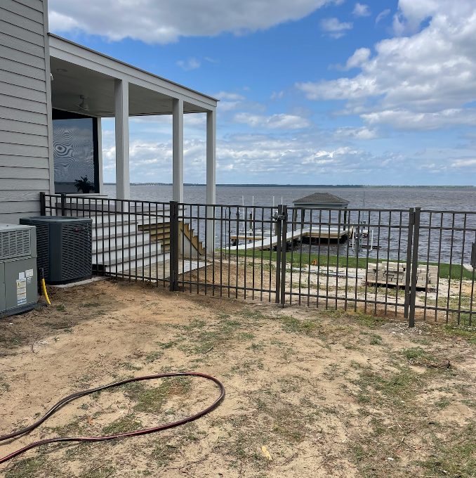 Black metal fence surrounds a sandy yard next to a house overlooking a body of water.