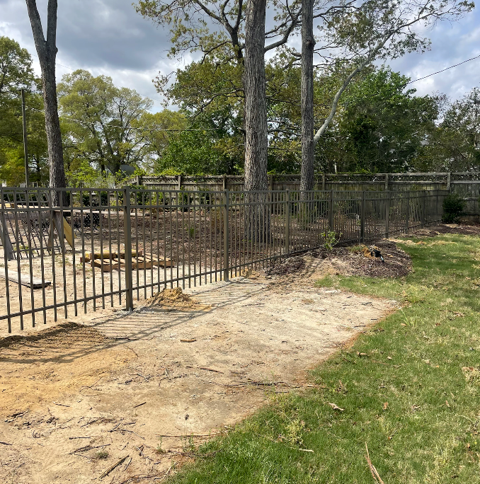 Metal fence encloses a sandy area between trees and a grassy lawn.