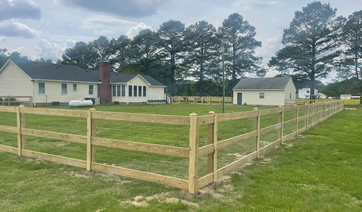 A two-rail wooden fence encloses a grassy yard with houses and trees in the background on a sunny day.