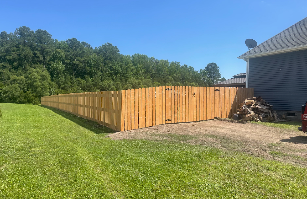 Wooden fence in a grassy backyard, with a house to the right and trees in the background.
