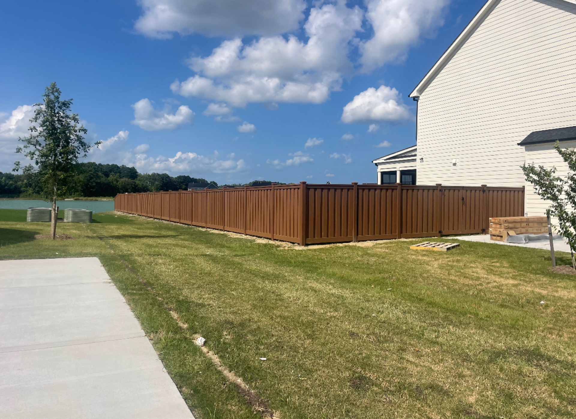 Brown wooden fence bordering a grassy yard and a house under a blue sky with clouds.