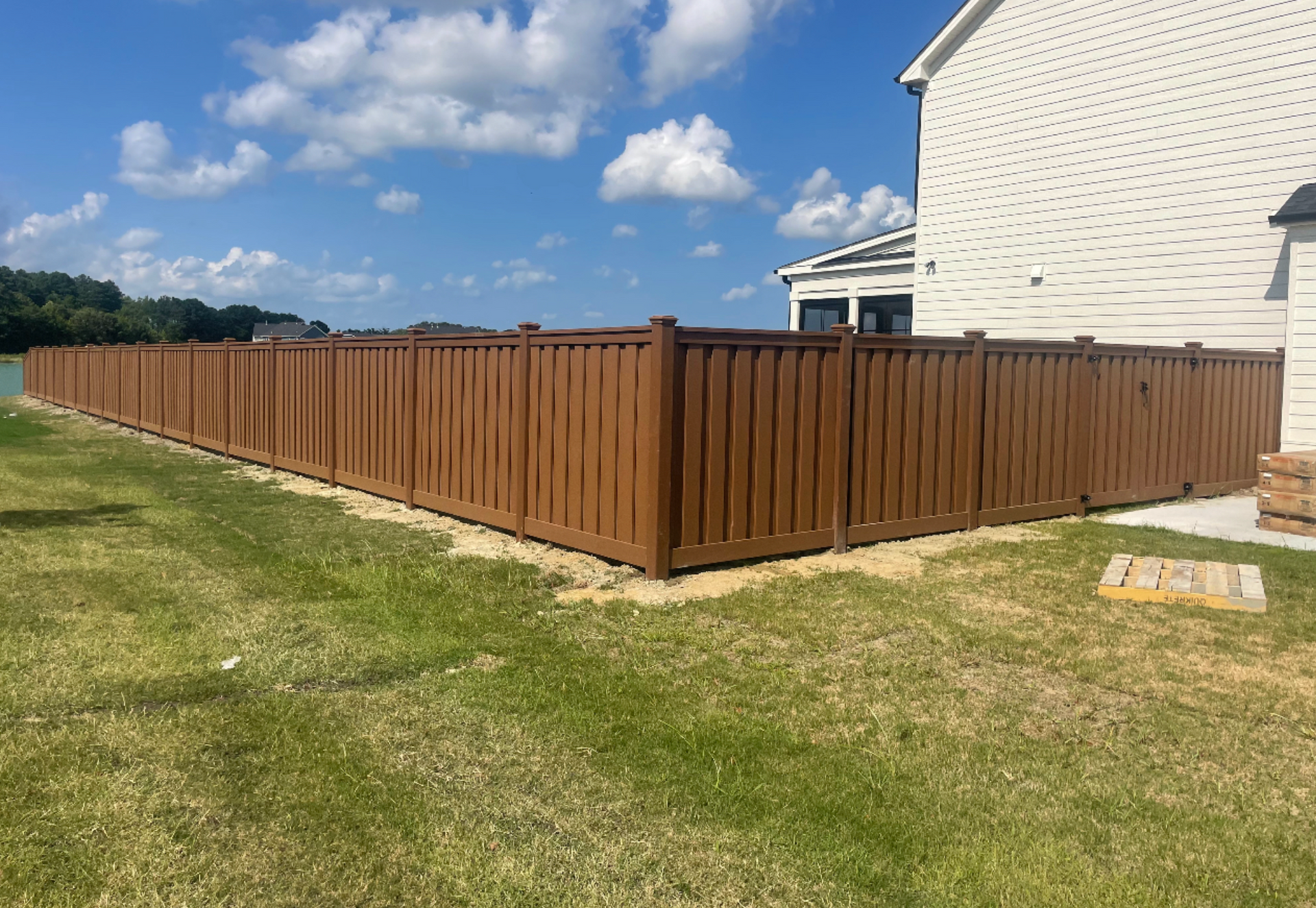 Brown wooden fence surrounding a grassy backyard, with a house in the background under a blue sky.
