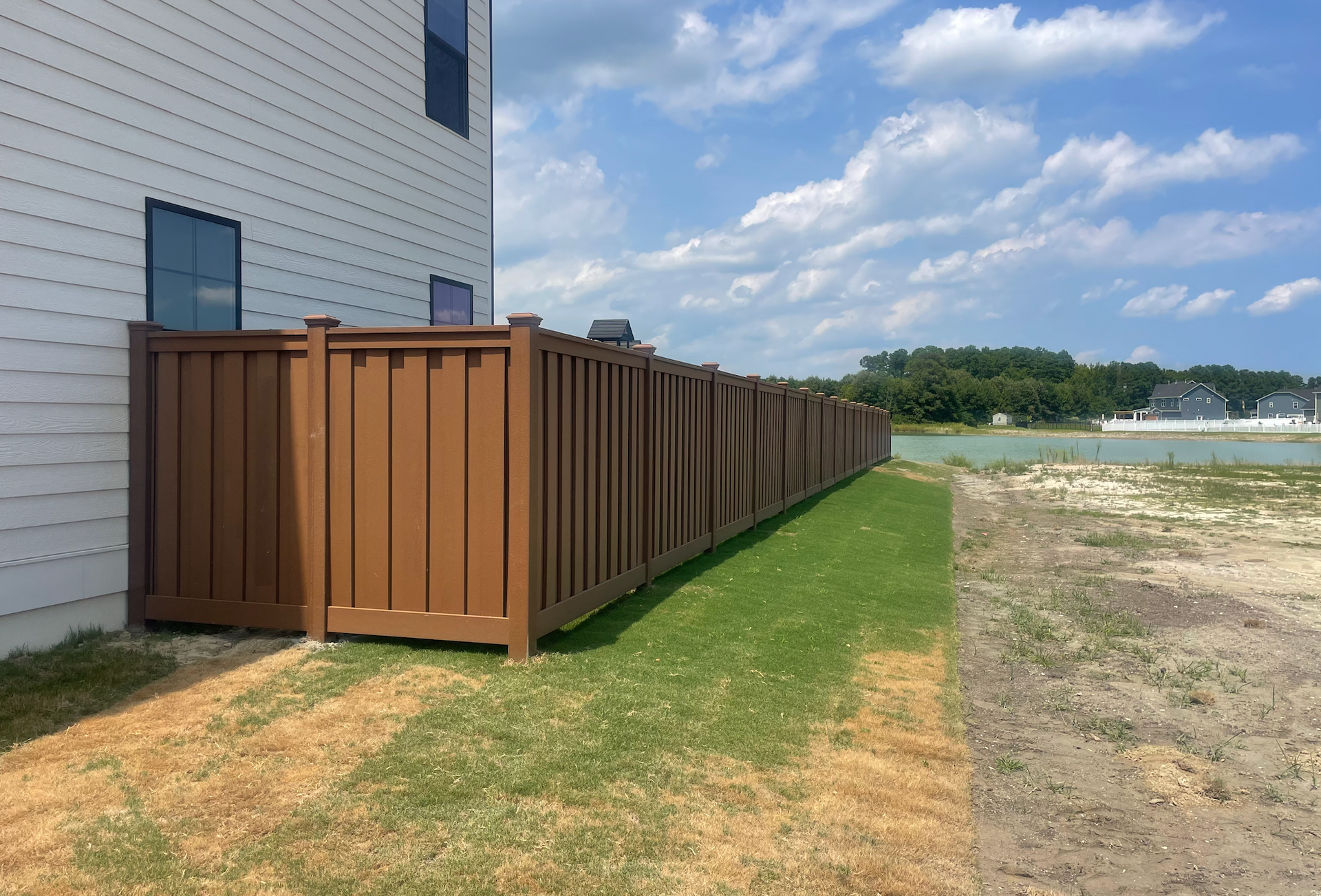 Brown privacy fence along the side of a white house, bordering a grassy area and a body of water.
