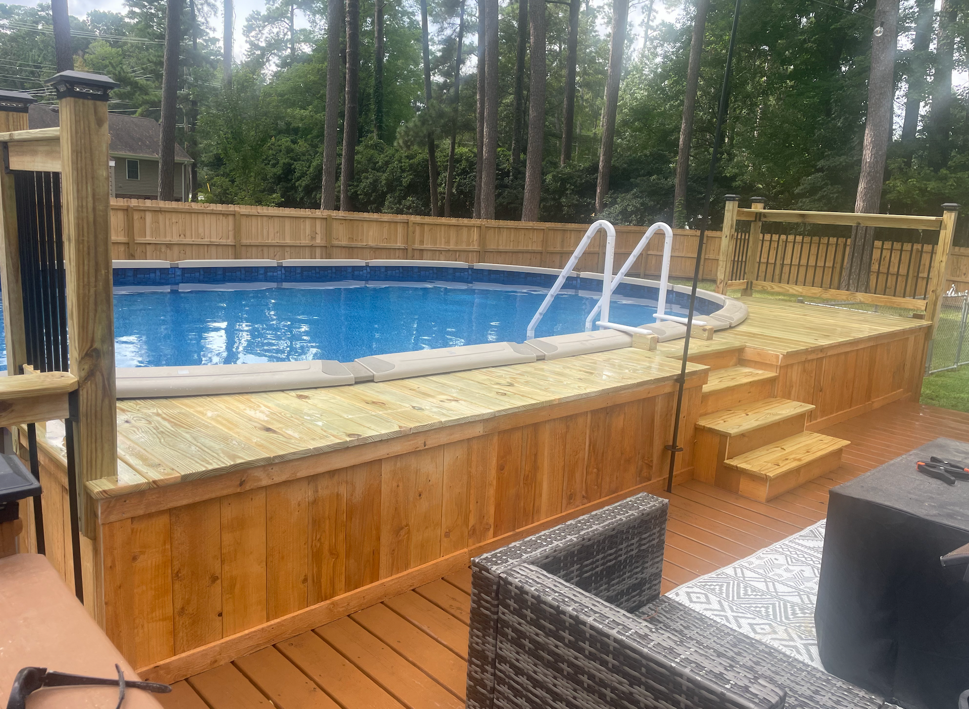 Above-ground pool with wooden deck, steps, and ladder. A patio with seating is in the foreground.