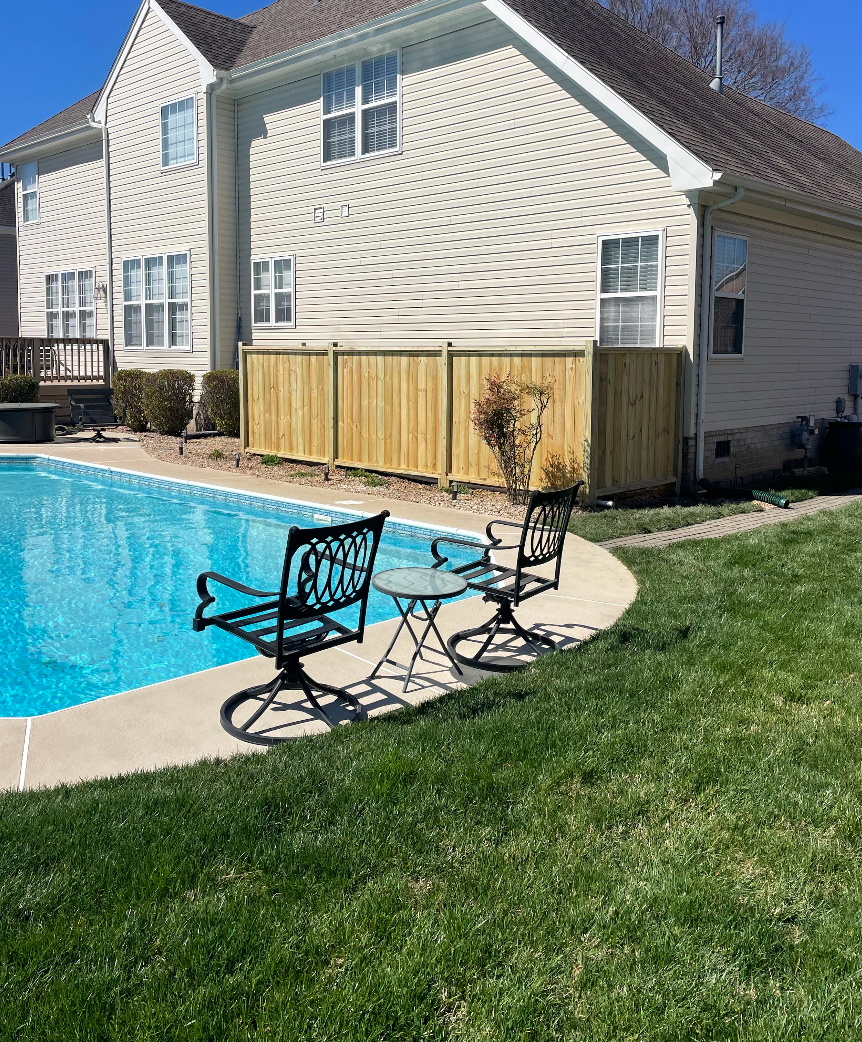 Two black chairs and a table sit by a pool with a wooden fence and a house in the background.