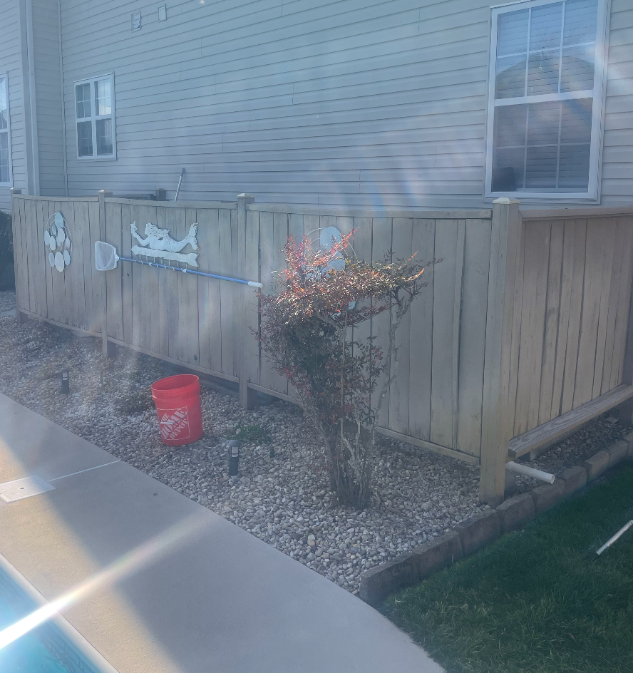 A wooden fence along a house with a red bucket, small plants, and decorative metal cutouts.