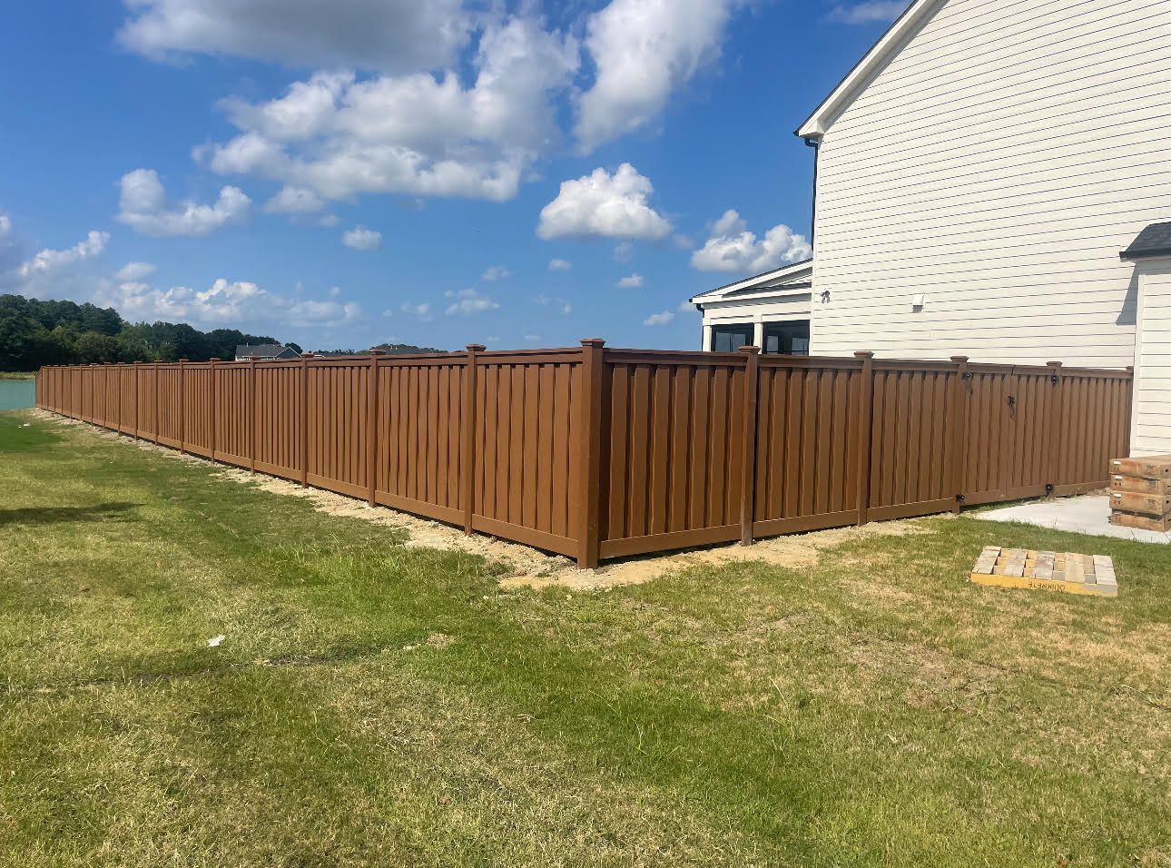 Brown wooden fence beside a white house on a grassy lawn under a blue sky