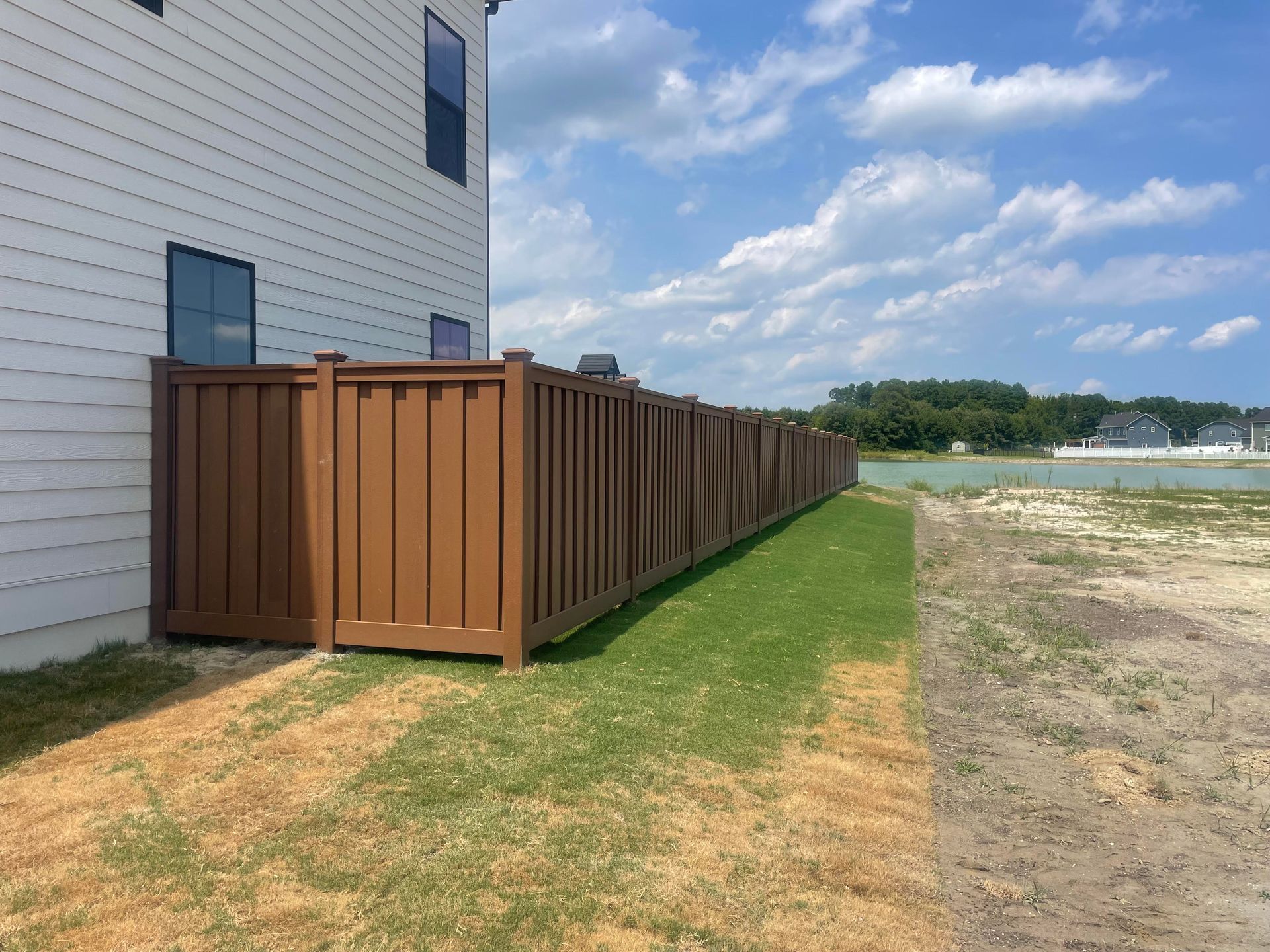Brown wooden privacy fence beside a white house, running along a grassy yard near water under a cloudy sky