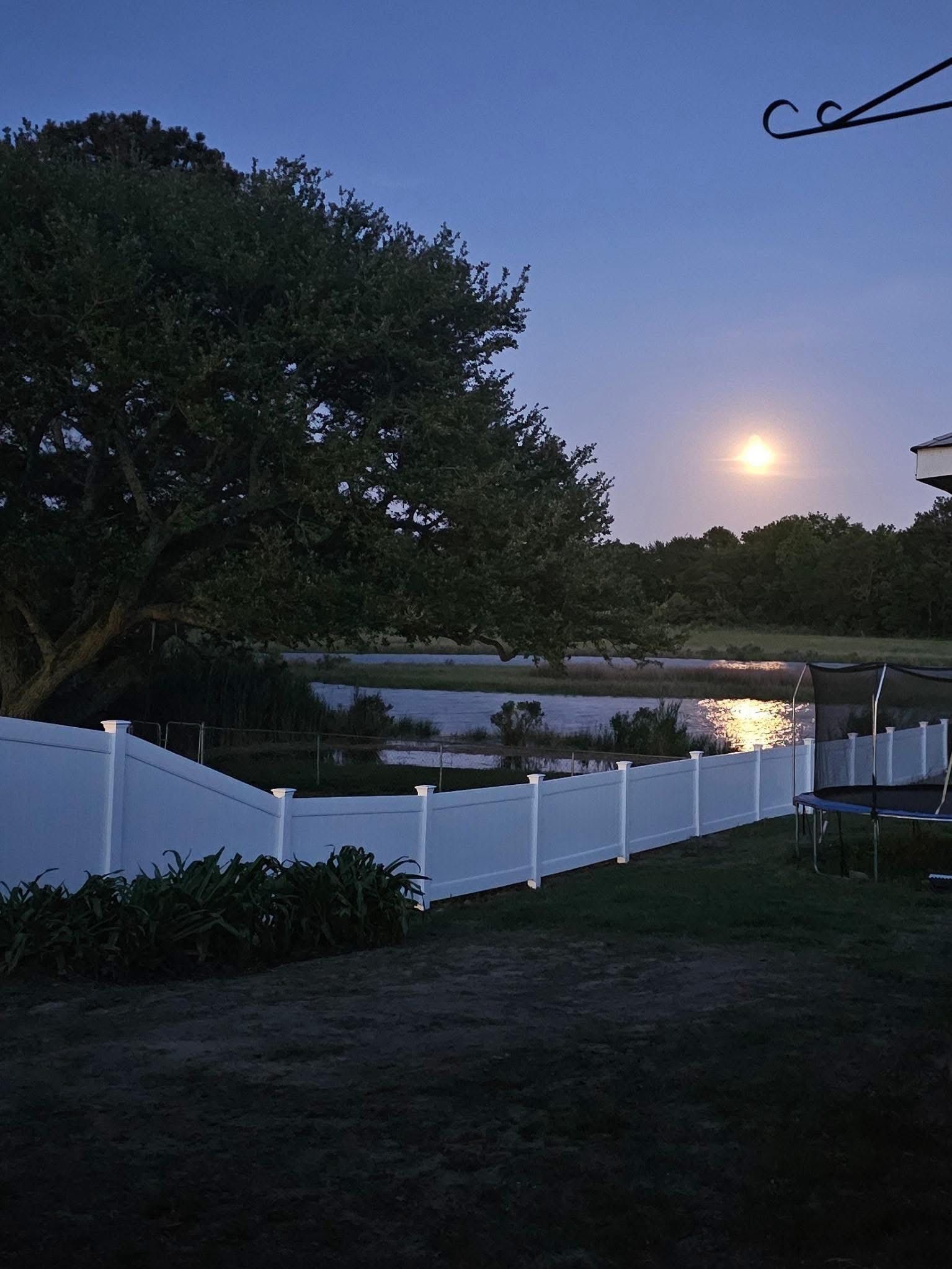 Dusk over a pond behind a white fence, with trees silhouetted and the moon glowing in the sky.