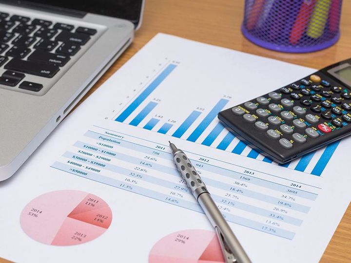 Spreadsheet with blue bar and pie charts, calculator, pen, and laptop on a desk