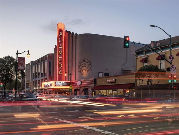 Neon-lit Alameda theater at dusk, with light trails from passing cars and a red traffic signal.