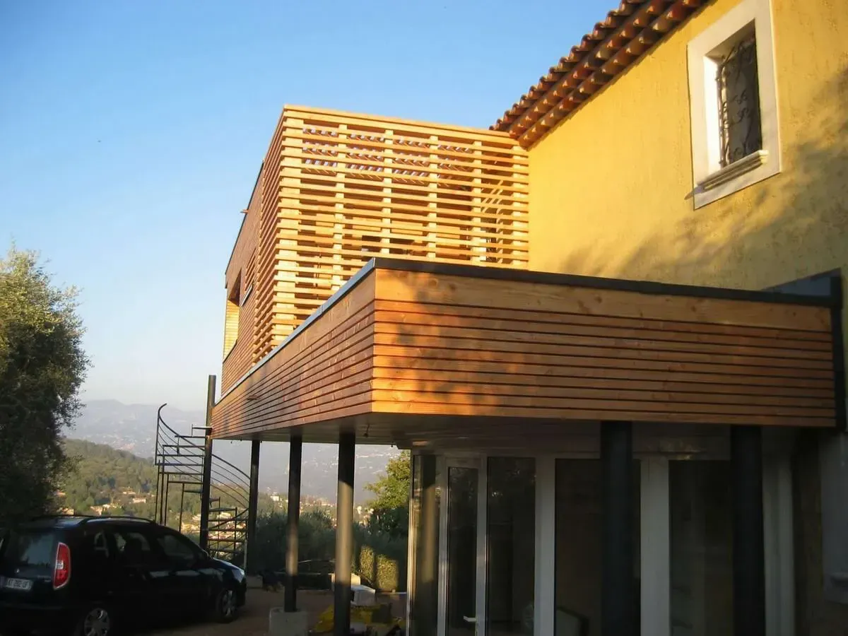 Extension moderne de maison avec murs à claire-voie en bois et balcon, soutenue par des piliers métalliques ; une voiture est garée à proximité.