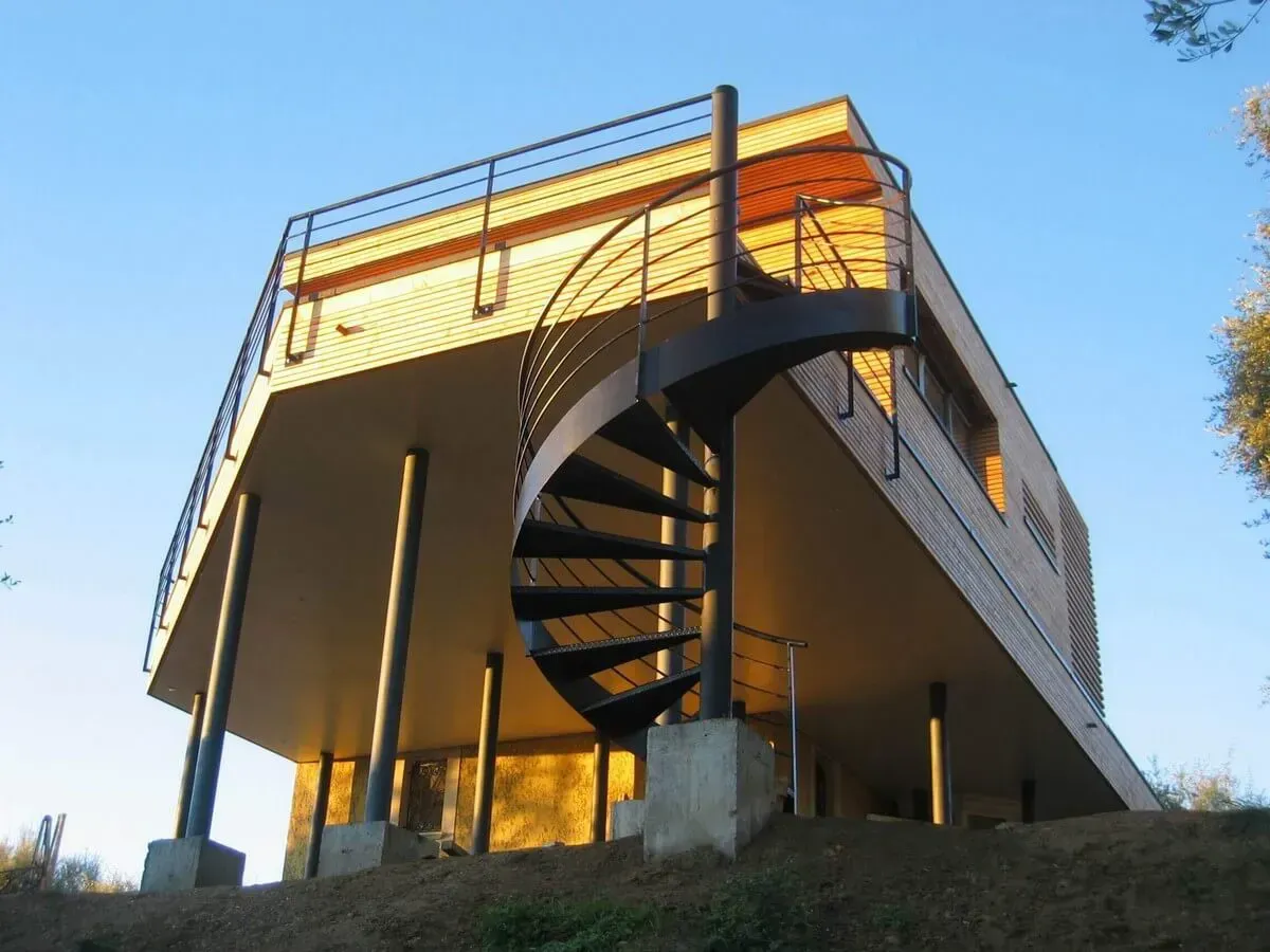 Maison moderne avec escalier en colimaçon noir ; vue extérieure. Bardage en bois, architecture surélevée, ciel bleu en arrière-plan.