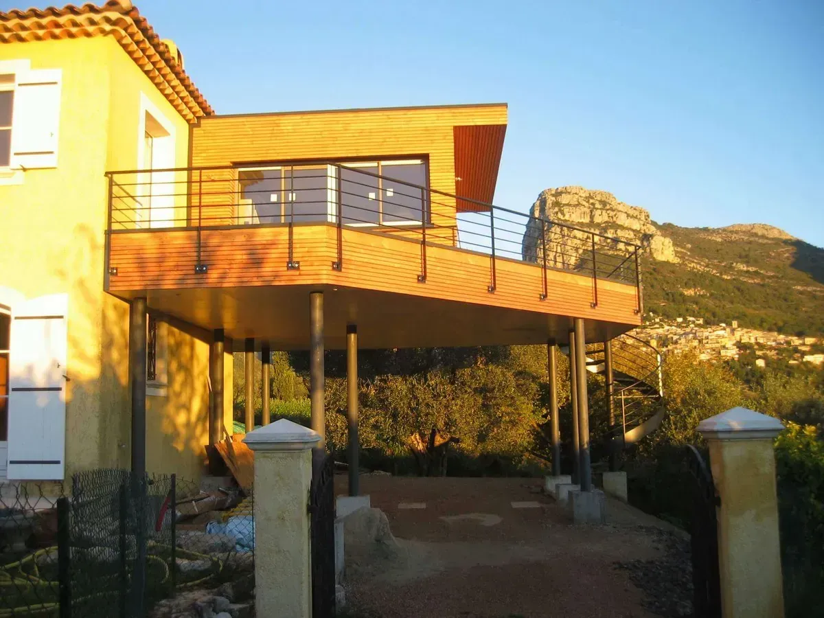 Maison à deux étages avec terrasse en bois, balustrades noires, soutenue par des piliers, avec la montagne en arrière-plan.