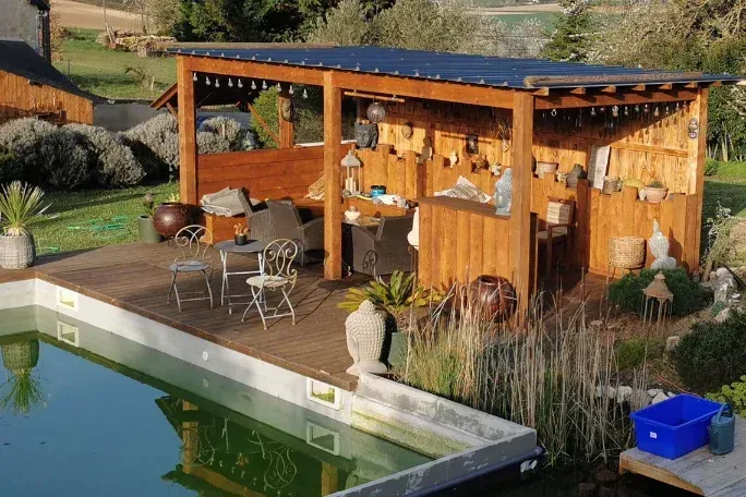Pergola en bois au bord de la piscine avec bar, chaises et aménagement paysager environnant.
