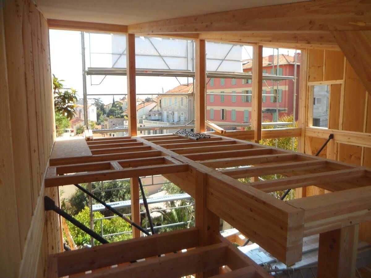 Vue intérieure d'un bâtiment en bois en construction, montrant la charpente du plancher, des murs et des ouvertures de fenêtres.