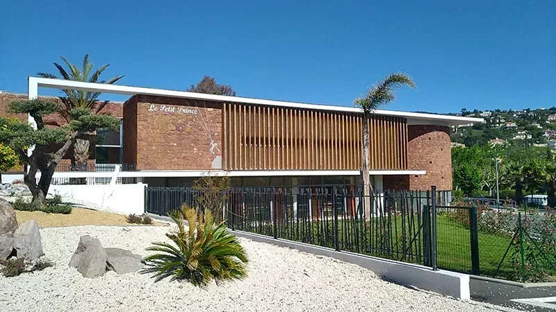 Bâtiment moderne en briques avec bardage en bois, palmiers et aménagement paysager en gravier blanc sous un ciel bleu.