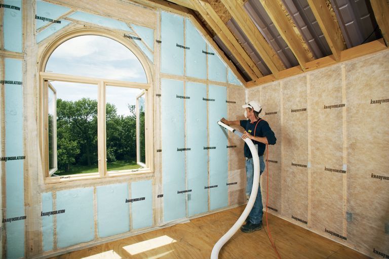 Workers on a roof installing trim; wooden house with large window, scaffolding, and evergreen tree against cloudy sky.