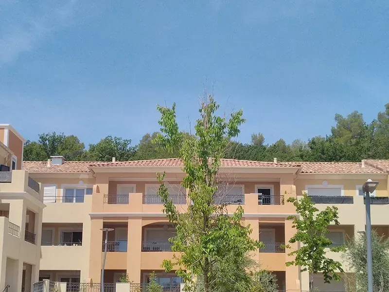 Immeubles d'appartements beiges aux toits de tuiles rouges, arbres au premier plan et ciel bleu clair.