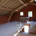 Intérieur d'un bâtiment en construction, avec poutres en bois apparentes, plancher partiellement construit et murs brun-rouge.