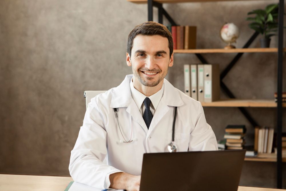 Médico sorridente sentado à frente de um notebook em consultório, transmitindo disposição e simpatia.