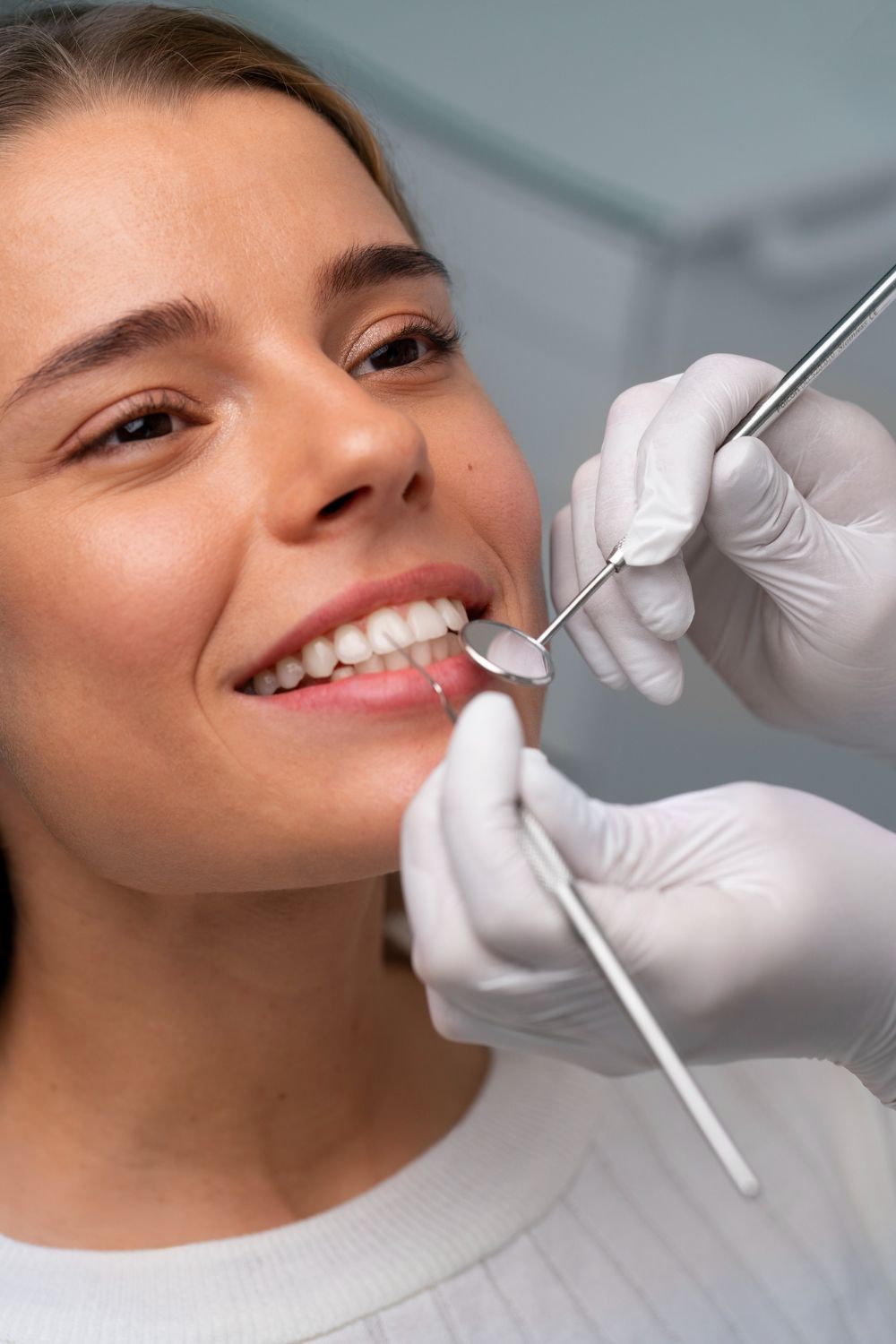 Close-up de uma mulher sorrindo enquanto um dentista realiza procedimento com espelho odontológico e instrumento de metal.
