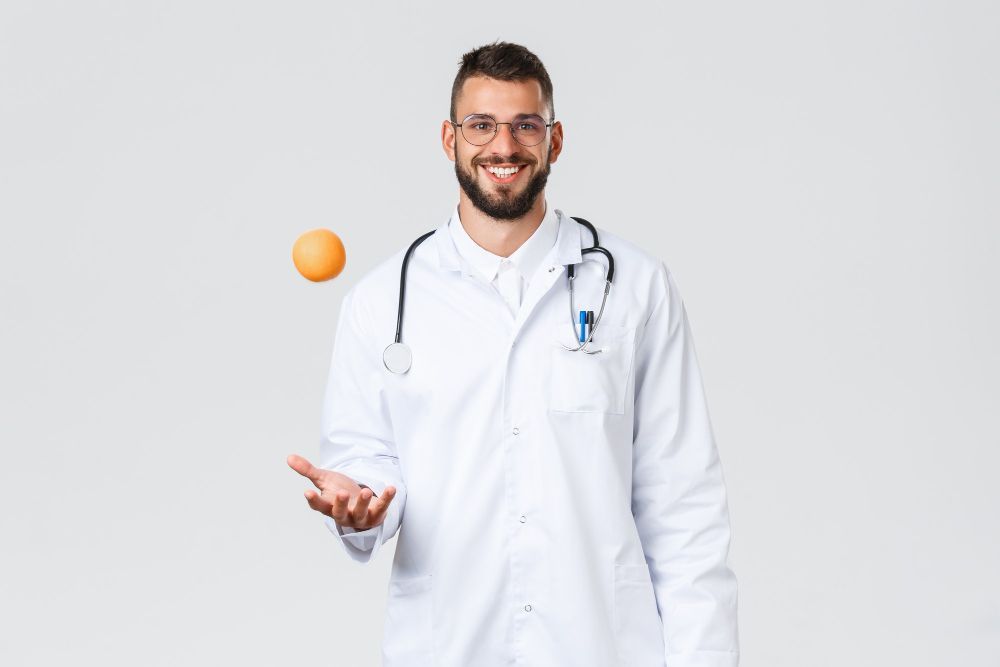médico sorridente com estetoscopio no pescoço jogando laranja para cima e fundo cinza