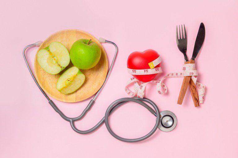 Prato com frutas, fita métrica, estetoscópio e talheres sobre fundo rosa, simbolizando alimentação saudável.