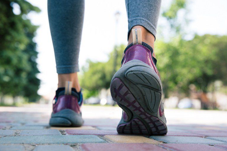 Pés de pessoa caminhando em calçada arborizada, representando caminhada como exercício físico.