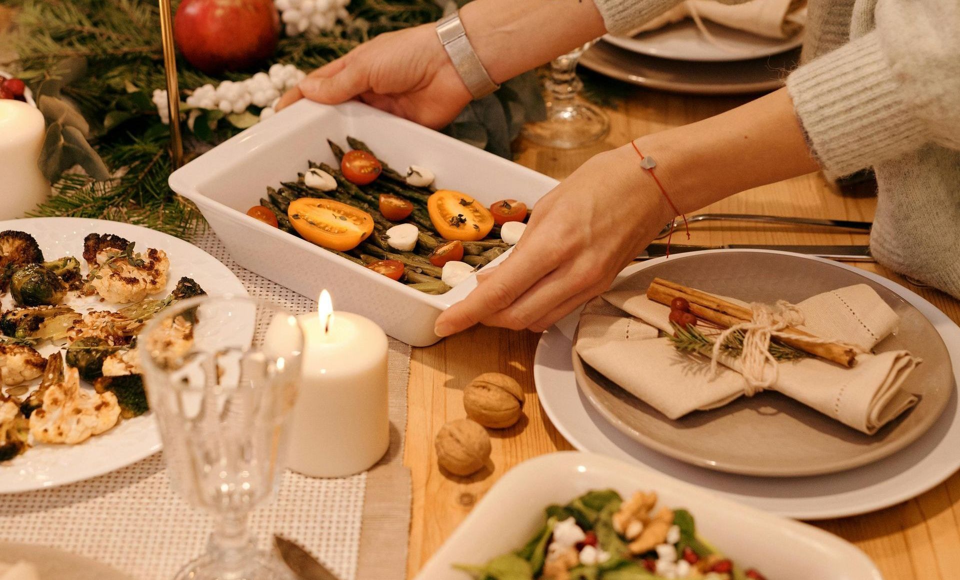 Mesa posta com pratos festivos, velas acesas e mãos servindo comida — imagem associada a festas de fim de ano.