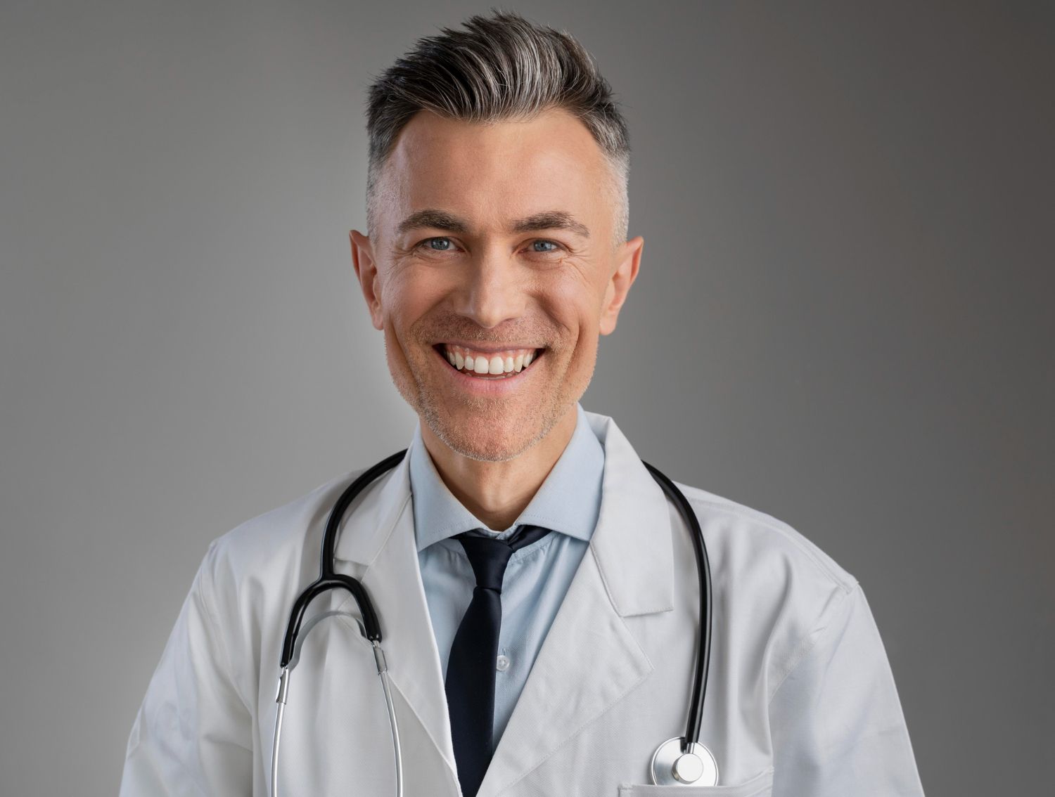 Médico sorridente de cabelos grisalhos usando jaleco e estetoscópio, posando em frente a fundo neutro.