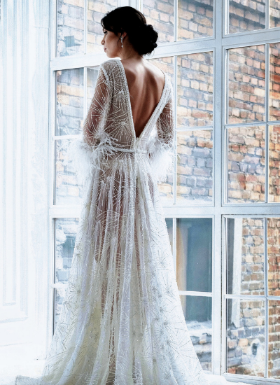 Woman in a white lace gown standing by a bright window, seen from behind.