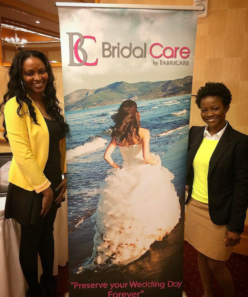 Two women posing beside a Bridal Care banner at an indoor event.