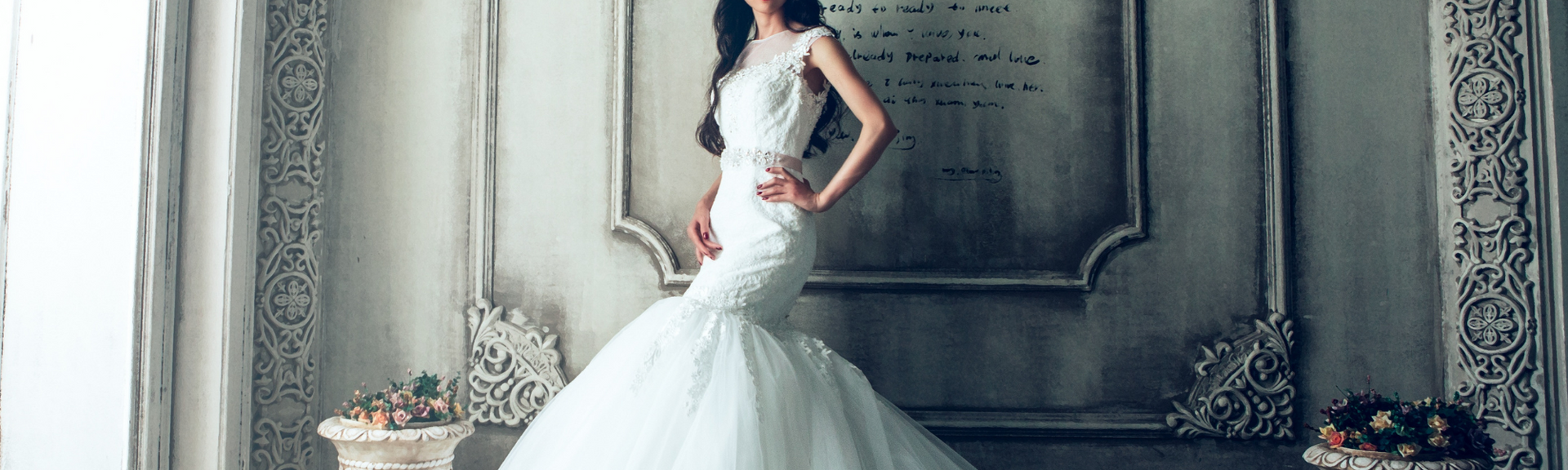 Woman in a white gown posing in an ornate gray interior, beside a floral arrangement.