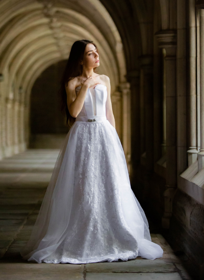 Woman in a white gown standing in a stone corridor with arched columns, lit by soft light.