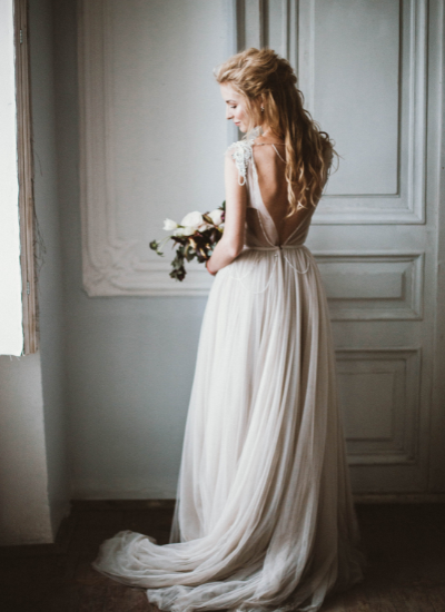 Woman in a flowing white gown holding flowers beside a window in a softly lit room