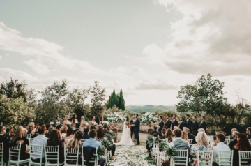 Outdoor wedding ceremony with guests seated on white chairs beside a floral aisle at sunset