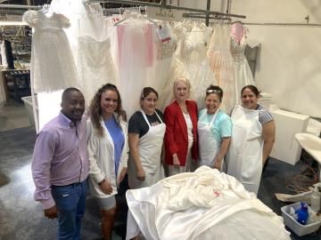 Group of six people standing in a bridal gown workshop, surrounded by hanging white dresses and fabric.