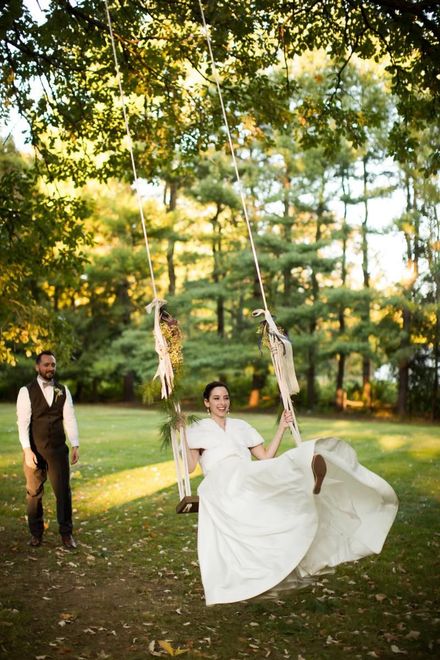 A bride swings on a rope swing in a sunlit park while a man stands nearby.