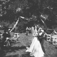 Bride in a white dress walks outdoors beneath a floral arch at a garden wedding ceremony