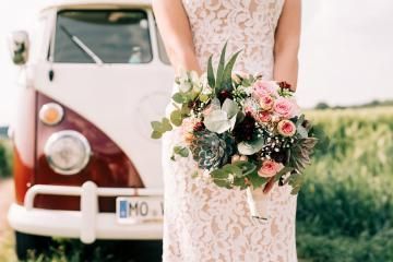 Bride holding a pink-and-white bouquet in front of a vintage van in a field