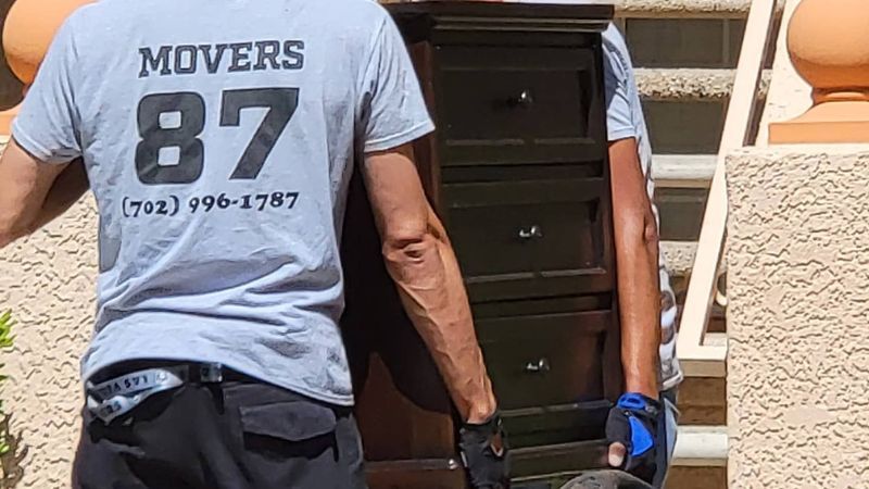 Two movers carry a dark dresser up outdoor stairs. One wears a shirt that says 