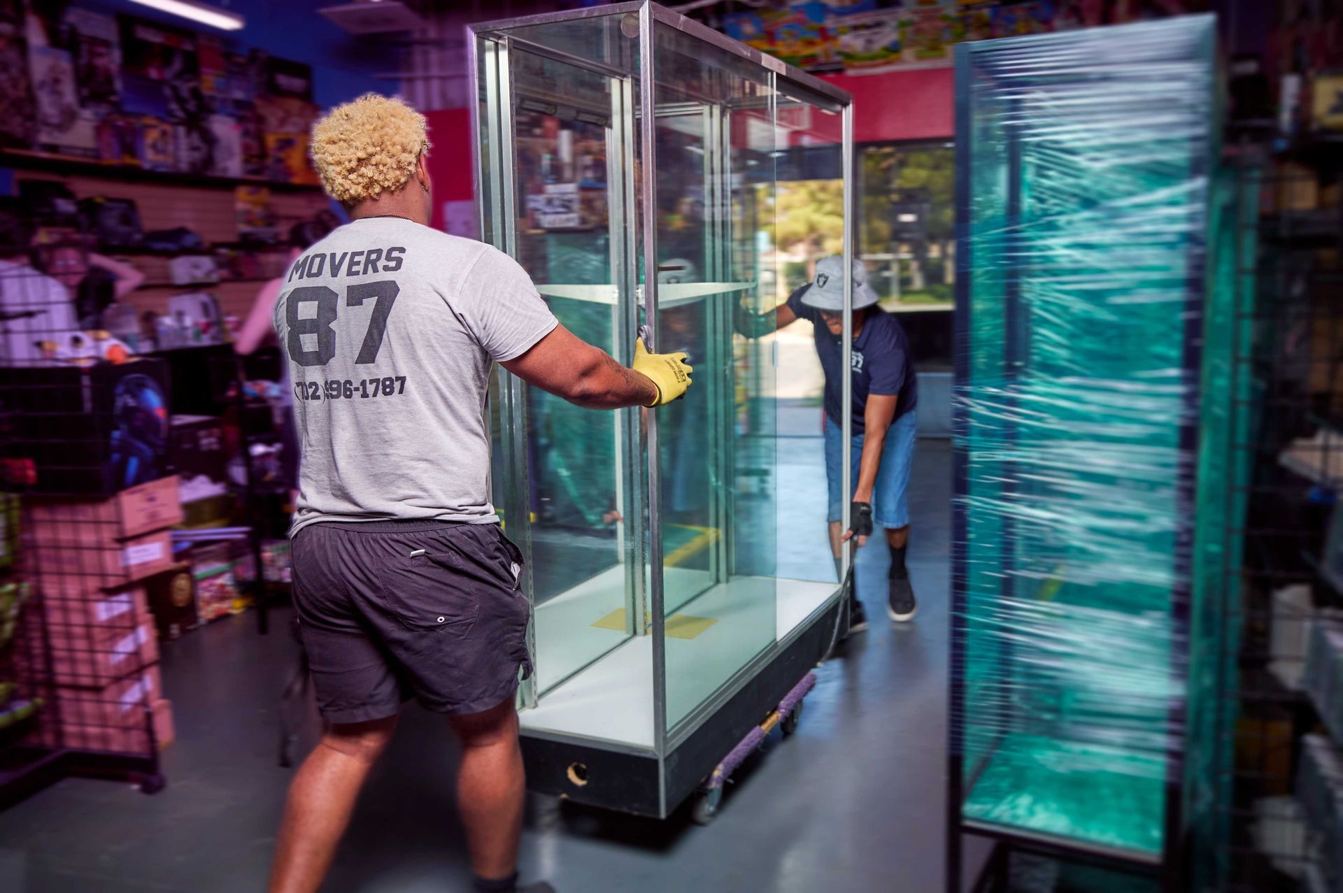 Two people moving a glass display case inside a store with shelves and merchandise.