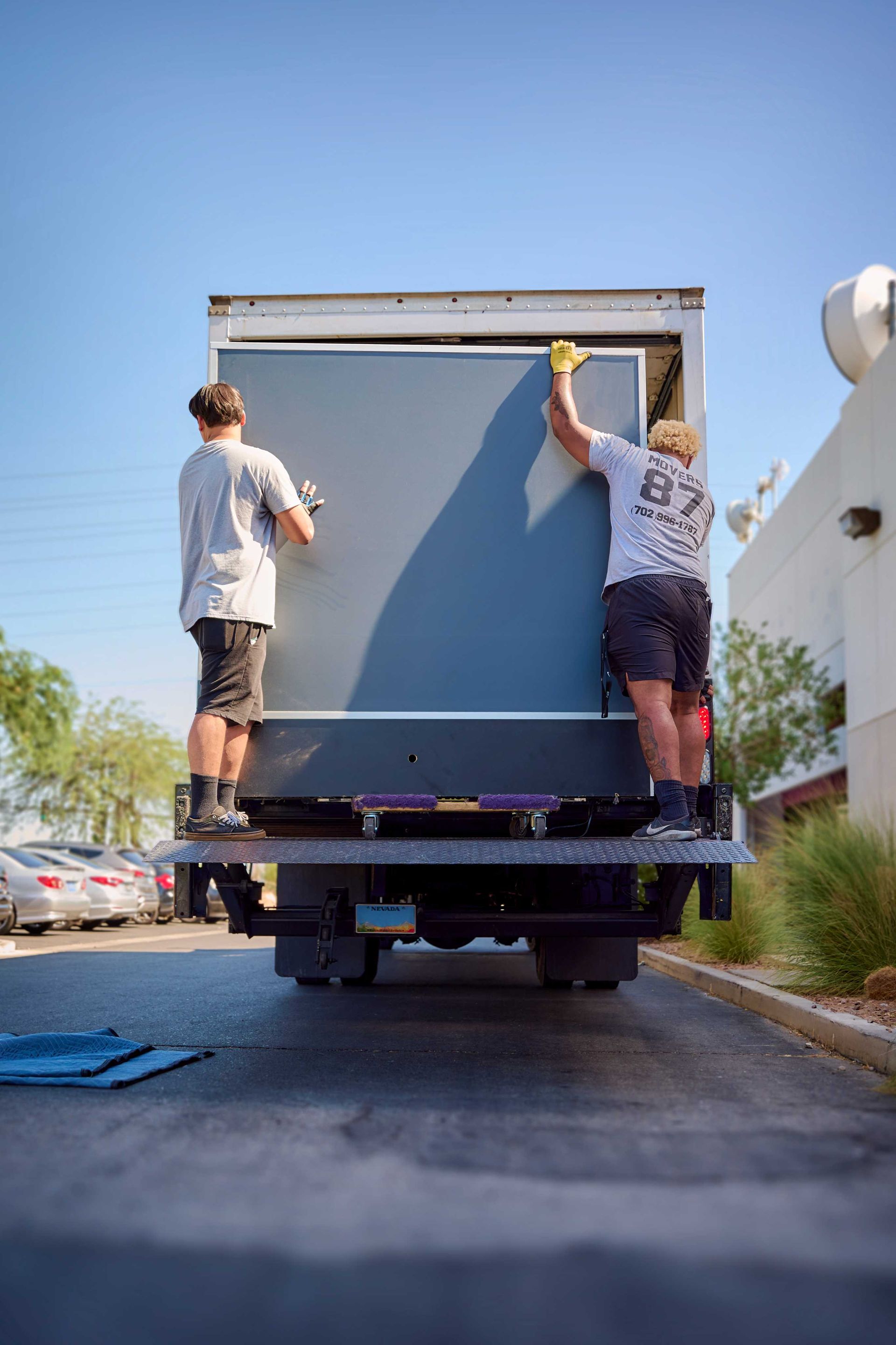 Two people loading a large gray object into the back of a box truck on a sunny day.