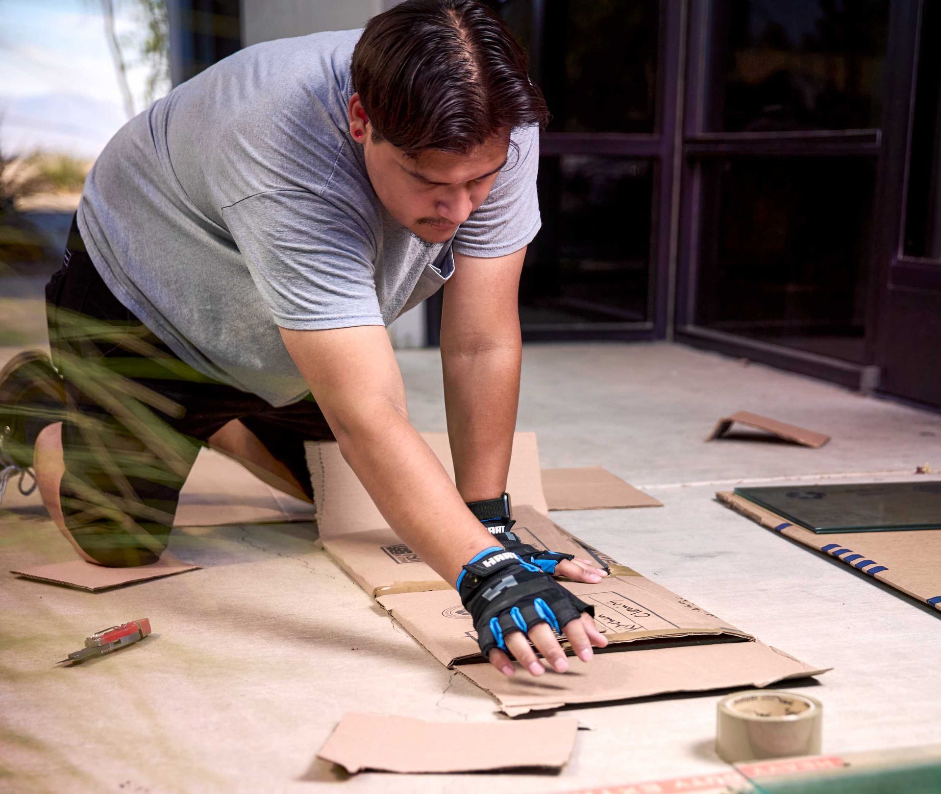 Man kneeling, cutting cardboard on a floor with a utility knife, wearing gloves.