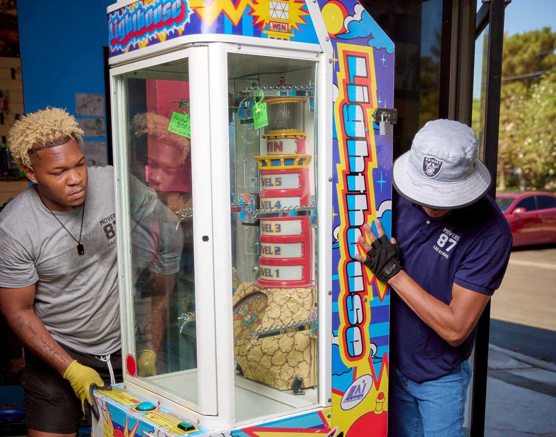 Two people move a claw machine outside a building. One wears a Raiders hat and the other has blonde hair.