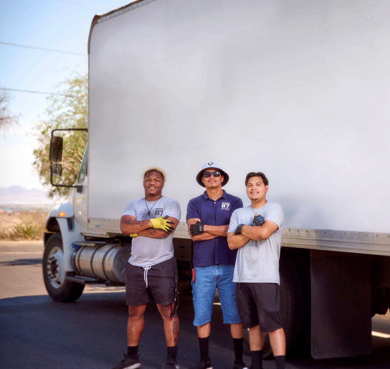 Three people standing in front of a moving truck with arms crossed, outside on a sunny day.